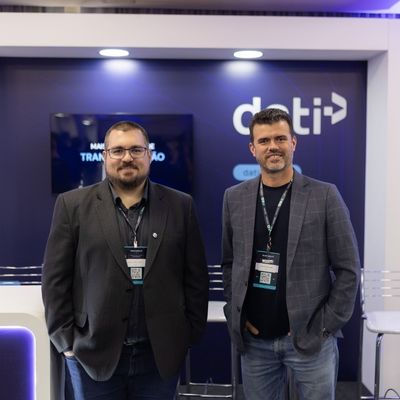 Dois homens usando blazers escuros e crachás de evento, em frente a um estande com o logo “DTI” no fundo.