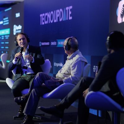 Três homens usando headsets participando de um painel no palco, com uma tela grande ao fundo exibindo a palavra TECNOUPDATE.