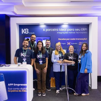 Grupo de seis pessoas sorrindo e posando em frente a um estande da K8 Fintech, com um fundo azul exibindo informações sobre produtos e parceiros.