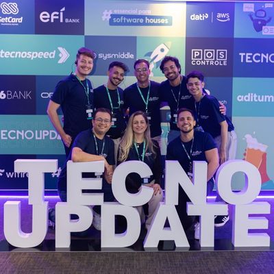 Sete pessoas sorrindo, usando camisetas pretas, posando atrás de grandes letras brancas formando a palavra "TECNO UPDATE" em um evento de tecnologia, com logos de patrocinadores ao fundo.