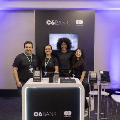 Quatro funcionários do C6 Bank sorrindo, em pé atrás de um estande com a marca e logos da Mastercard, em um ambiente de evento interno.