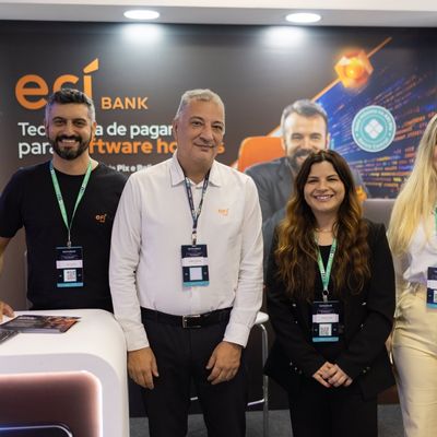 Três profissionais sorrindo, usando crachás de evento, em frente a um painel promocional do EFÍ Bank.