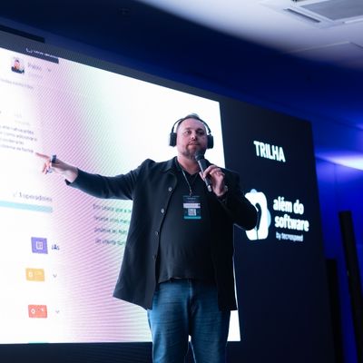 Apresentador usando headset e microfone, gesticulando em frente a uma tela grande com interface de software e identidade visual do evento.