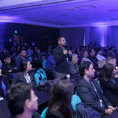 Homem em pé, falando ao microfone durante uma conferência, com o público sentado ao redor em um ambiente com pouca luz e iluminação azul.