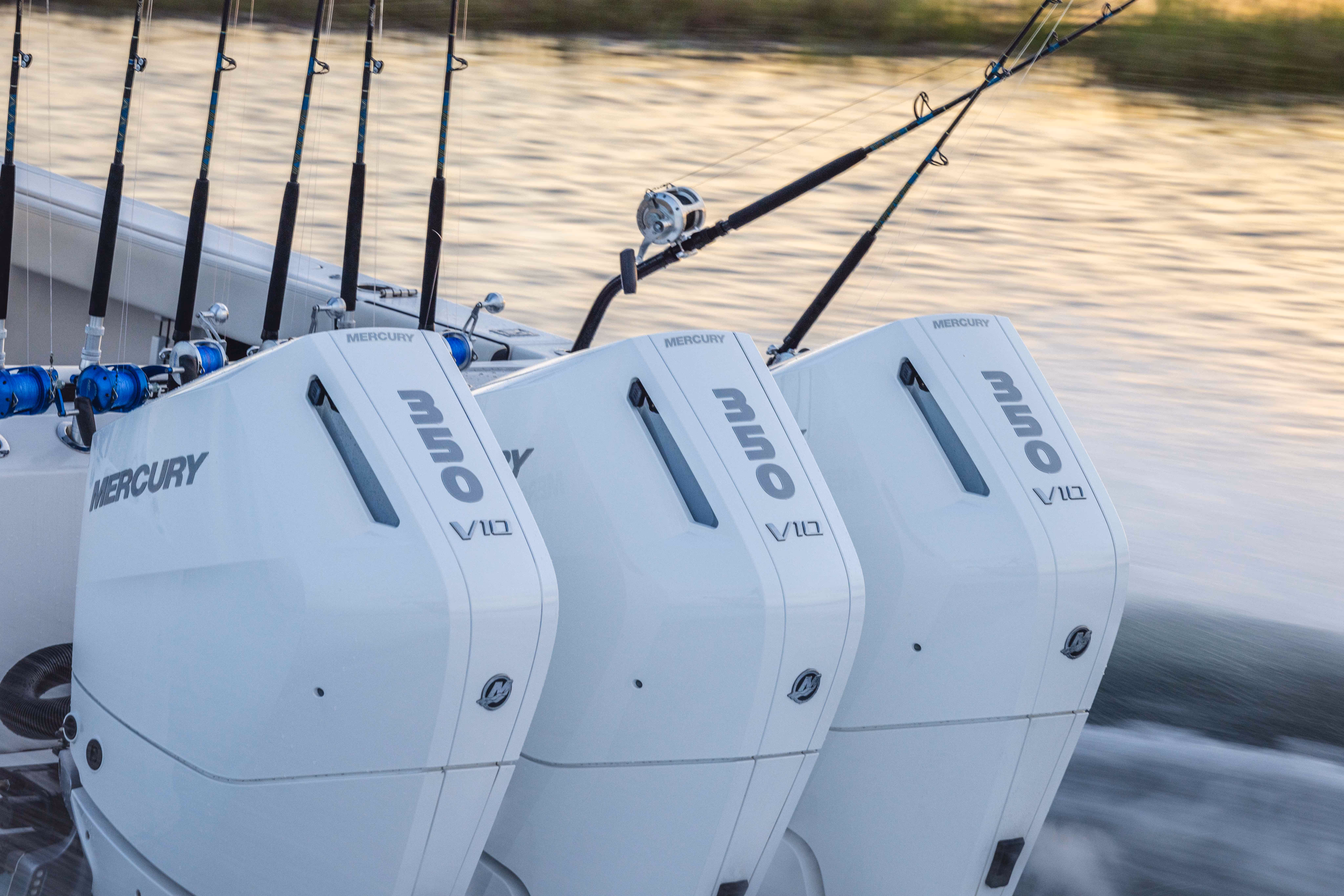 Three white Mercury 350 V10 outboard boat engines mounted side by side on a boat with fishing rods in the background on water.