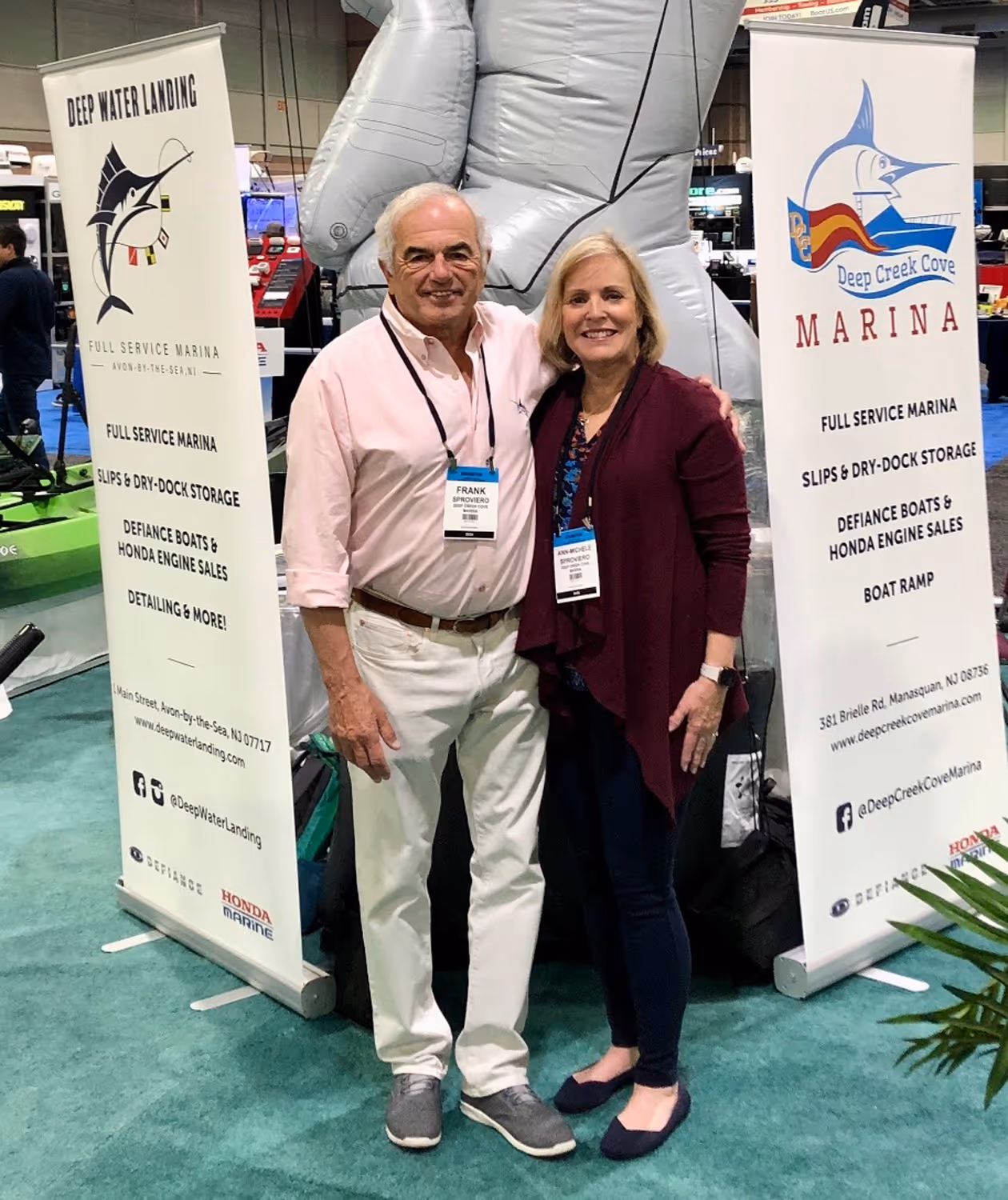 A man and a woman standing and smiling in front of two marina advertising banners at an indoor event.