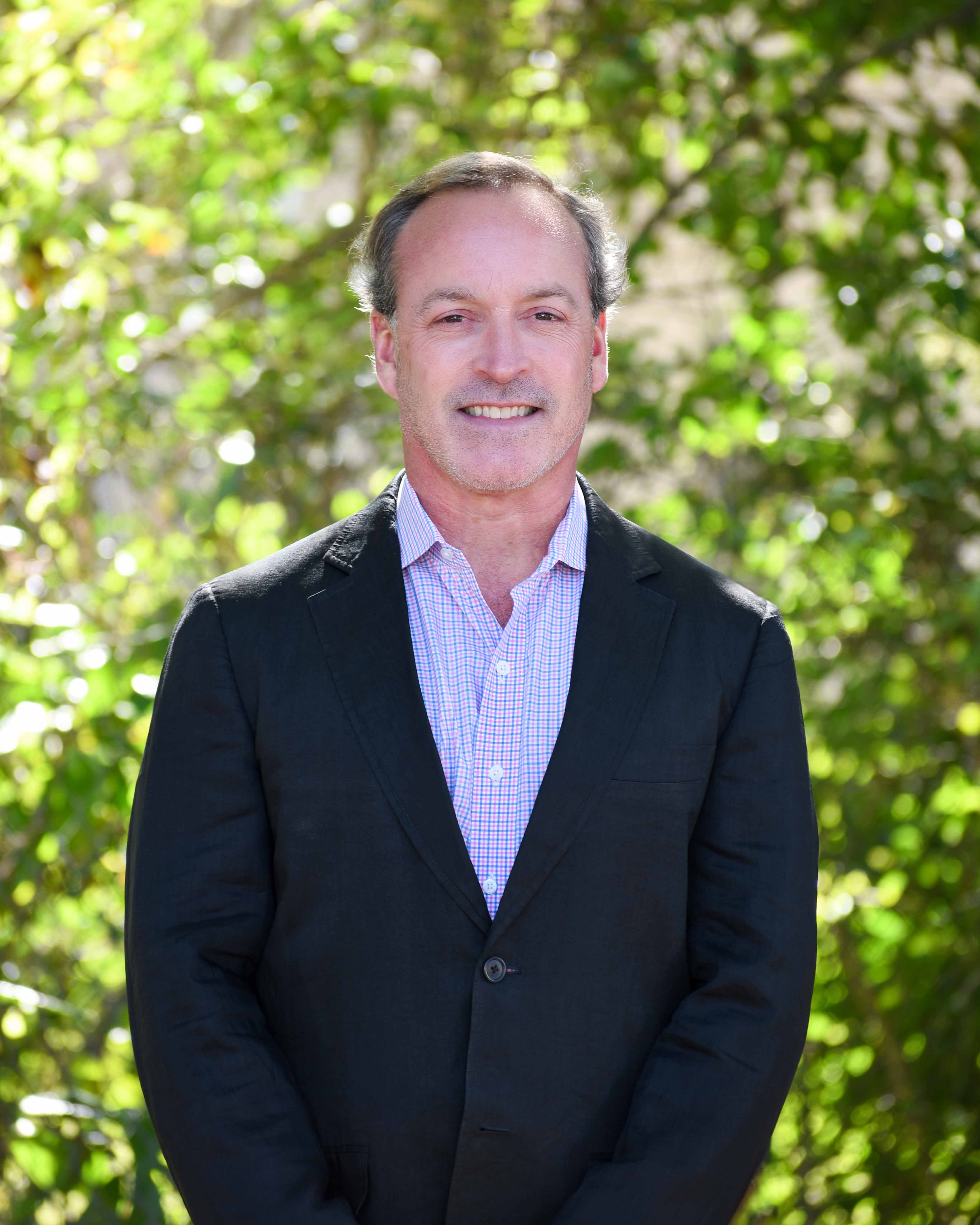 Smiling middle-aged man in a black blazer and checkered shirt standing outdoors in front of green foliage.