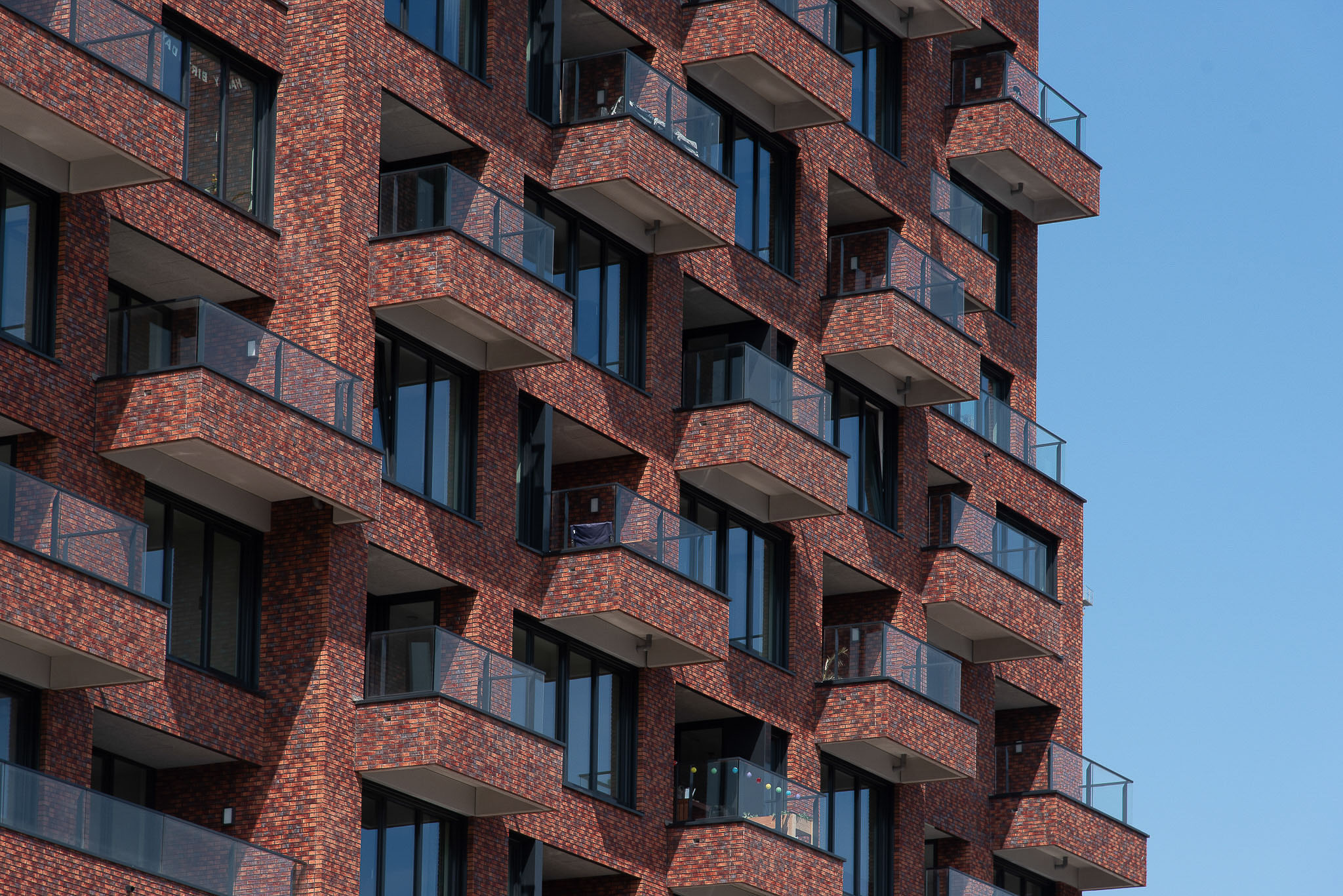 Close-up of a modern brick apartment building with multiple balconies featuring glass railings against a clear blue sky.