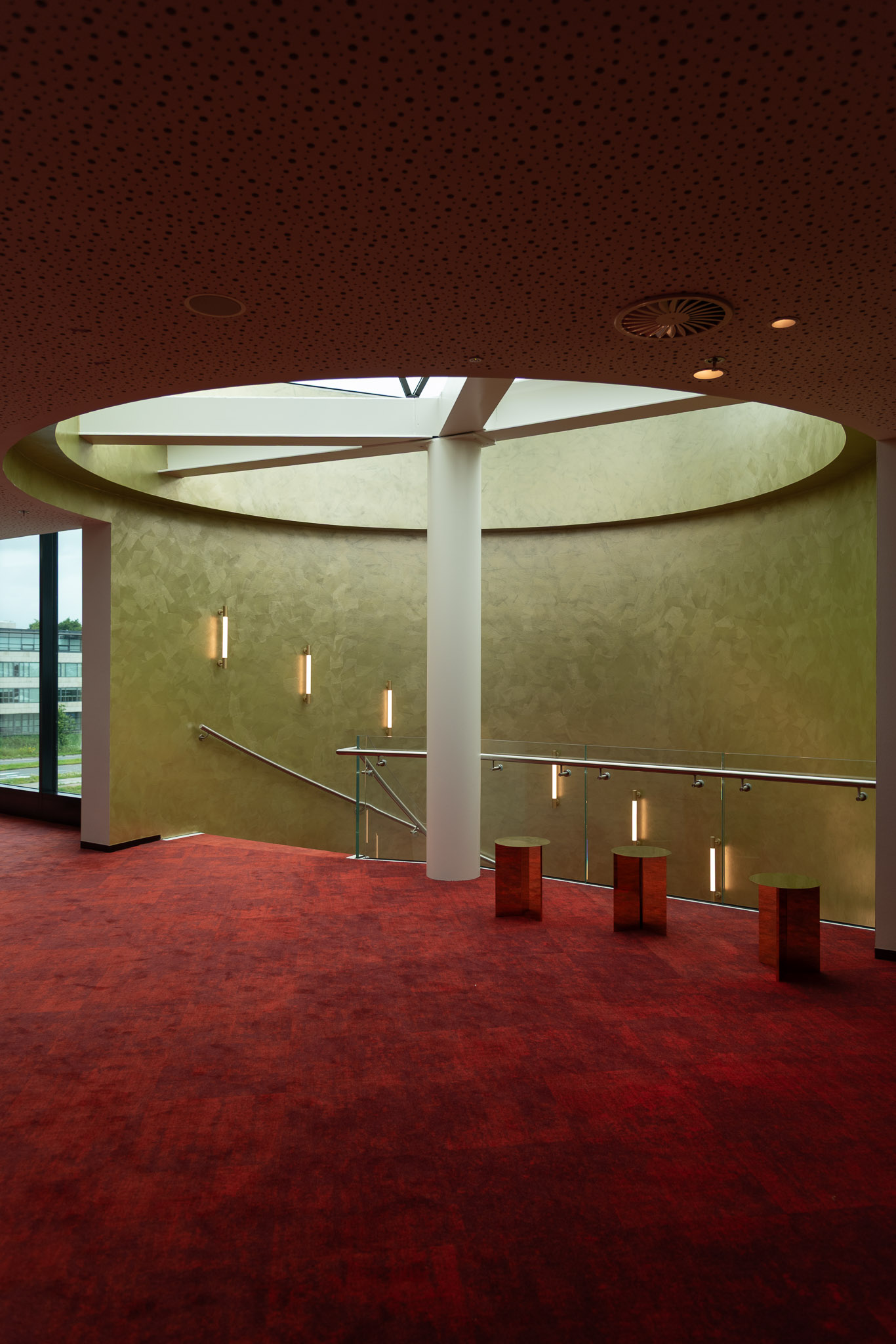 Interior view of a modern staircase area with red carpet, green textured curved wall, vertical light fixtures, and three cylindrical stools under a circular ceiling opening.