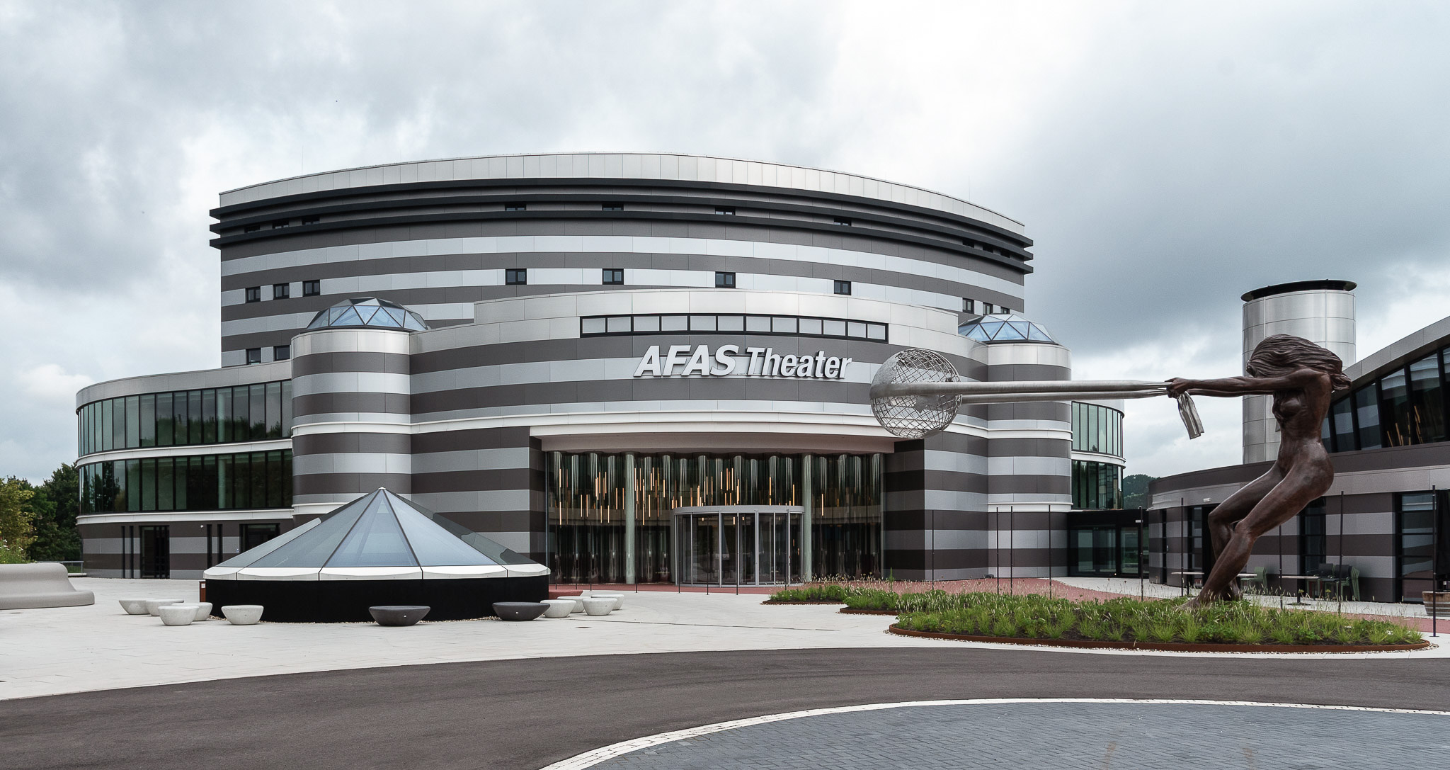 Modern AFAS Theater building with curved striped facade, glass entrances, and large bronze sculpture of a woman holding a globe.