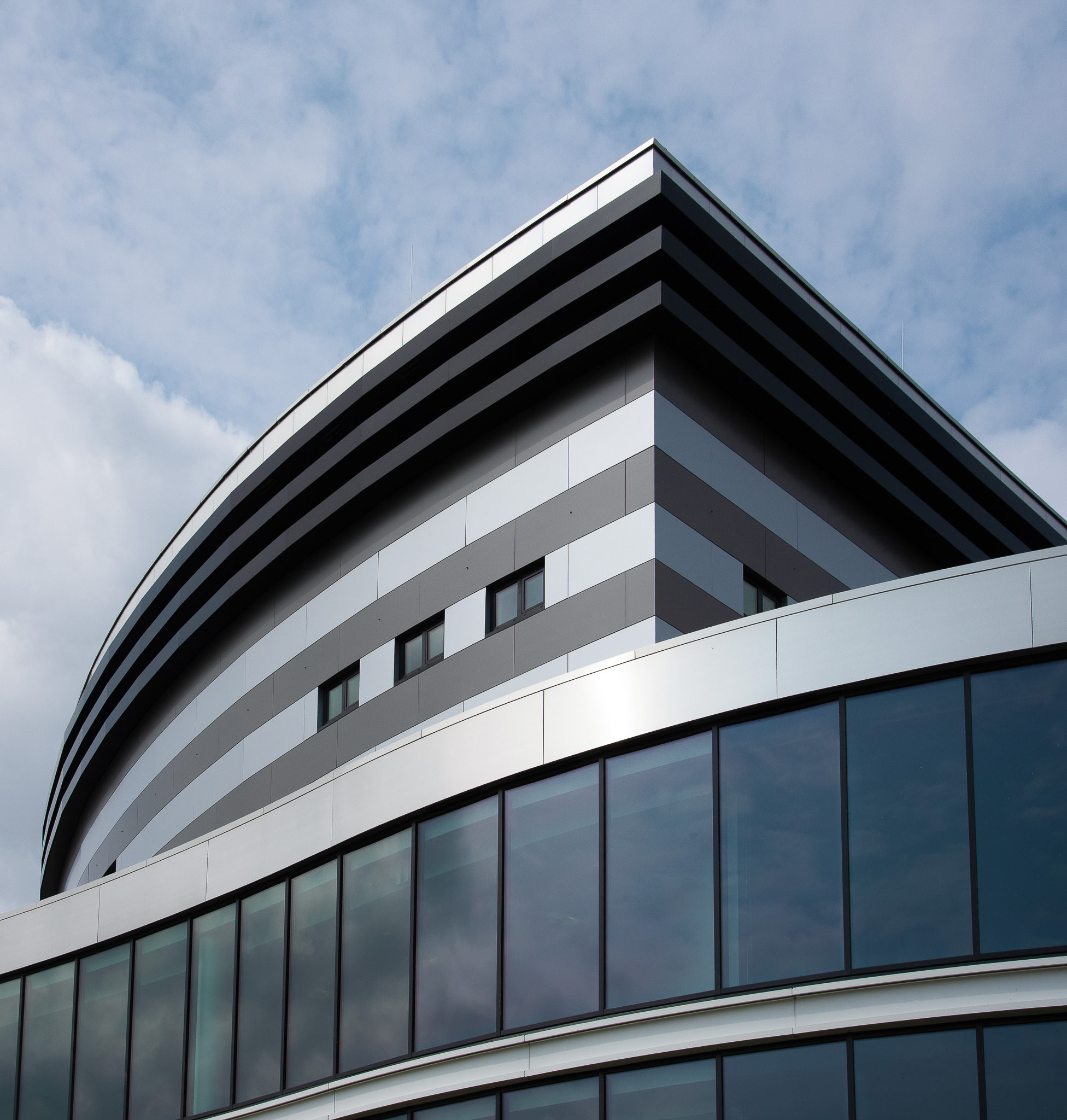 Modern architectural building with curved glass windows and striped gray facade under a partly cloudy sky.