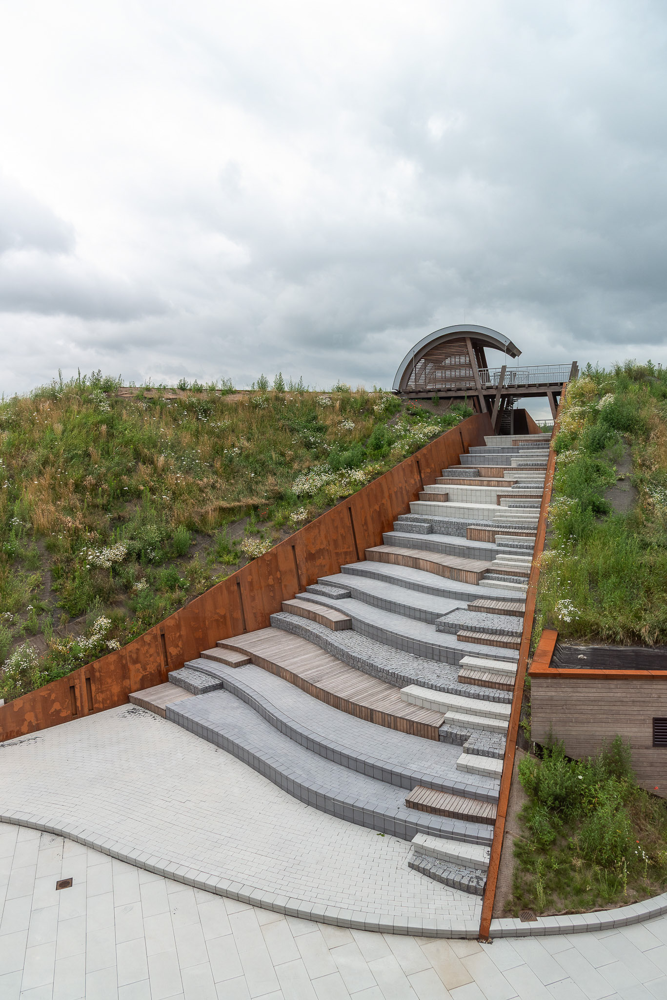 Stepped outdoor seating area with mixed materials integrated into a grassy hillside under a cloudy sky.