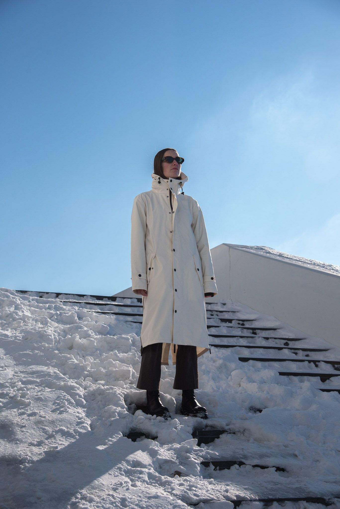 Person wearing a long beige coat and sunglasses standing on snowy steps under a clear blue sky.