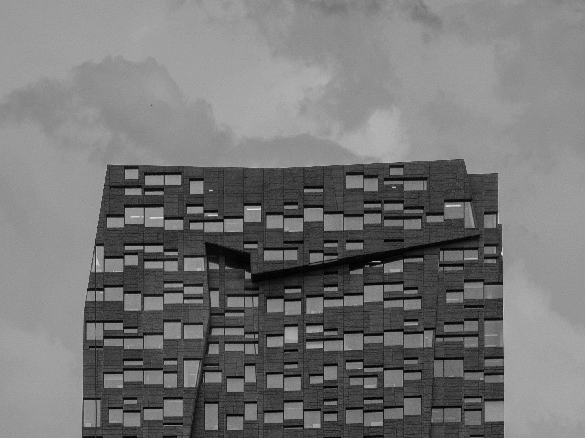 Modern high-rise building facade with an irregular pattern of rectangular windows and angular architectural features under a cloudy sky.