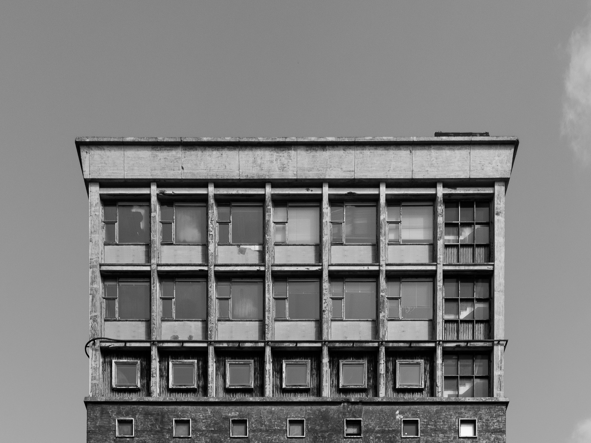 Black and white image of the top floors of an old, weathered multi-story building with various square and rectangular windows.