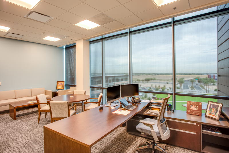 Commercial office workspace with large windows, roller shades, and natural light overlooking an exterior landscape.