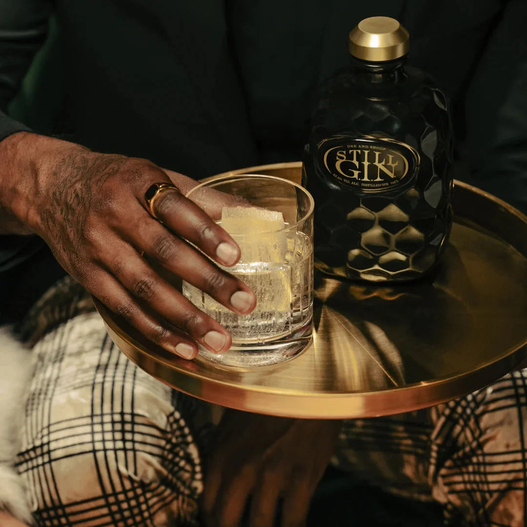Person lifting a cocktail glass from a brass tray beside a faceted black Still GIN bottle