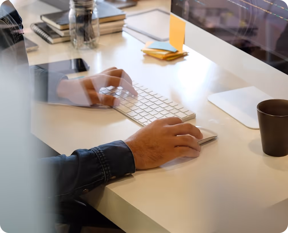 Person typing on a white keyboard and using a computer mouse on a desk with notebooks, sticky notes, and a coffee cup nearby.