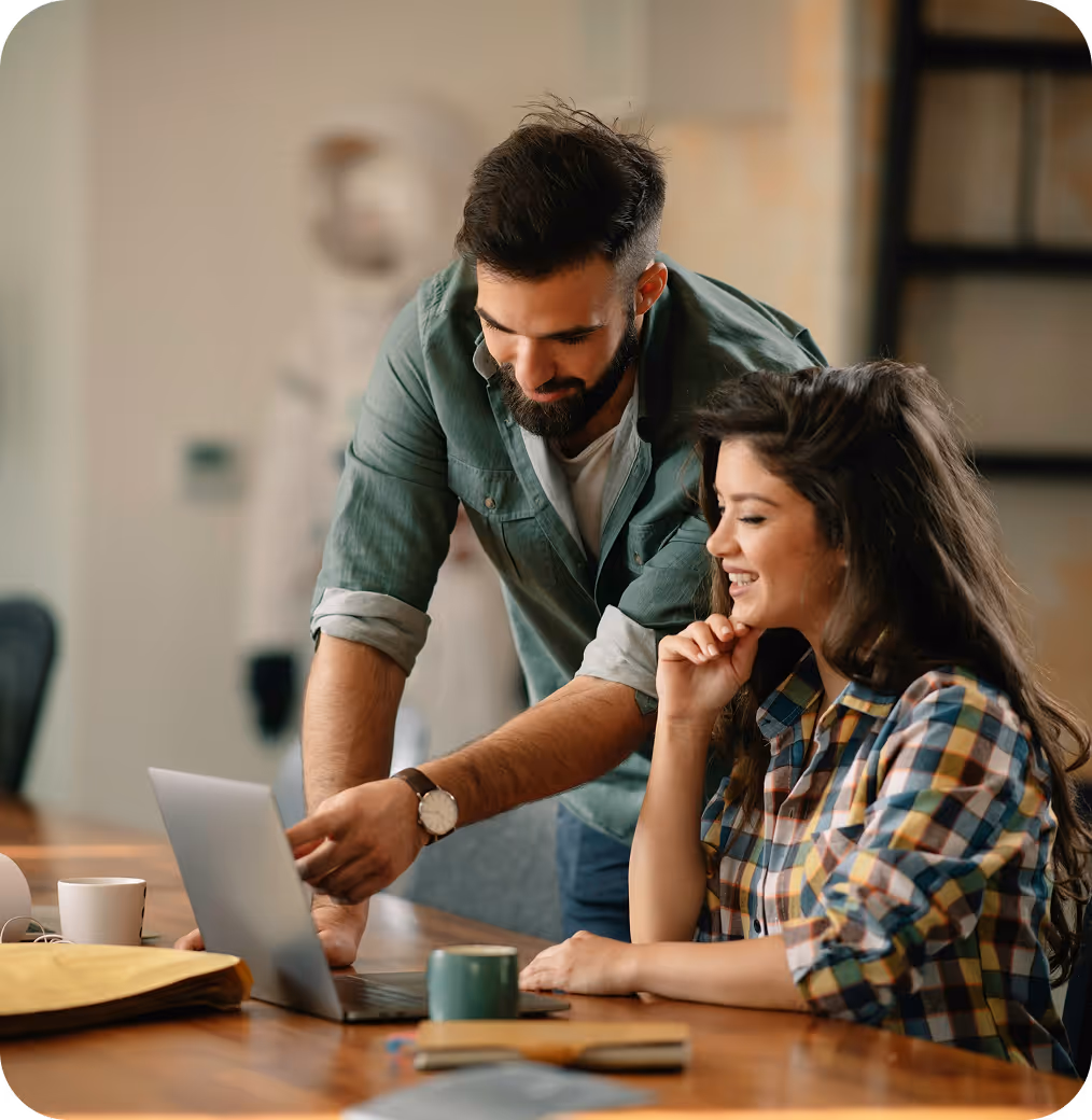 A man and a woman working together on a laptop at a wooden table, both smiling and engaged.