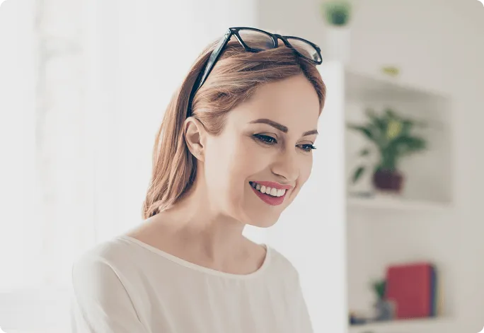 Smiling woman with glasses resting on her head working or reading indoors with a blurred background.