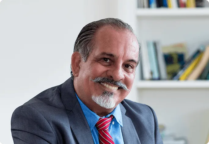 Smiling senior businessman with a mustache and goatee wearing a suit, blue shirt, and red striped tie in an office setting.