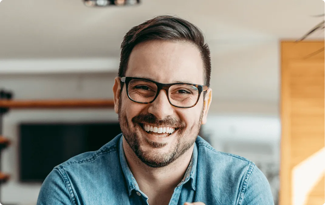 Smiling man with glasses and short beard wearing a blue denim shirt indoors.