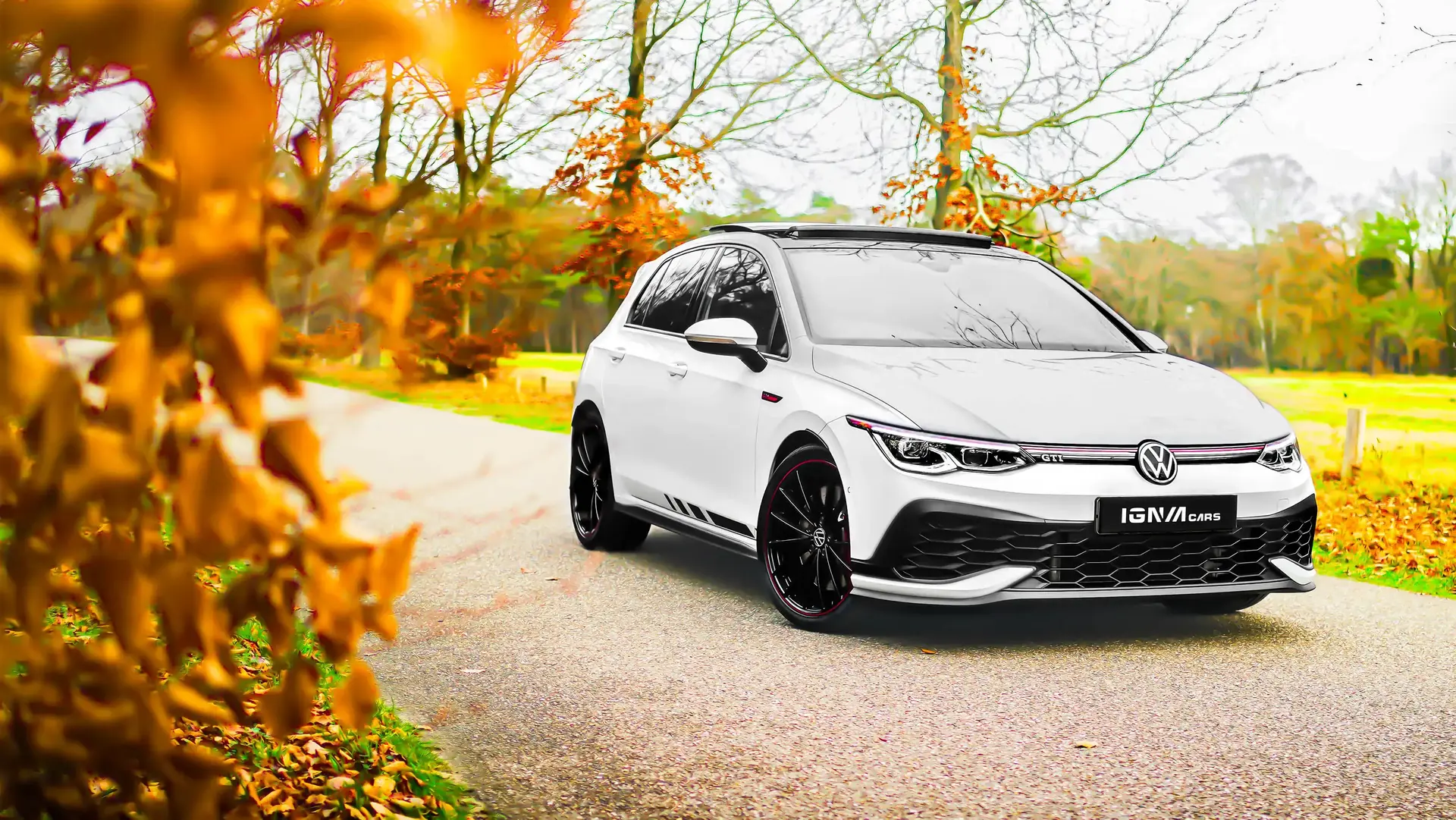 Witte Volkswagen GTI met zwarte velgen op een landweg omgeven door herfstbomen.