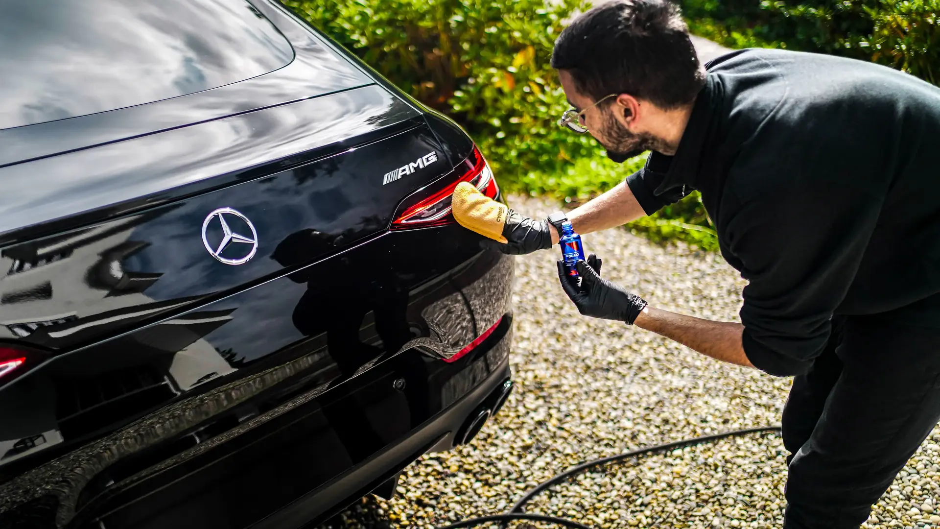 Man met handschoen brengt vloeistof aan op de achterkant van een glanzende zwarte Mercedes AMG.