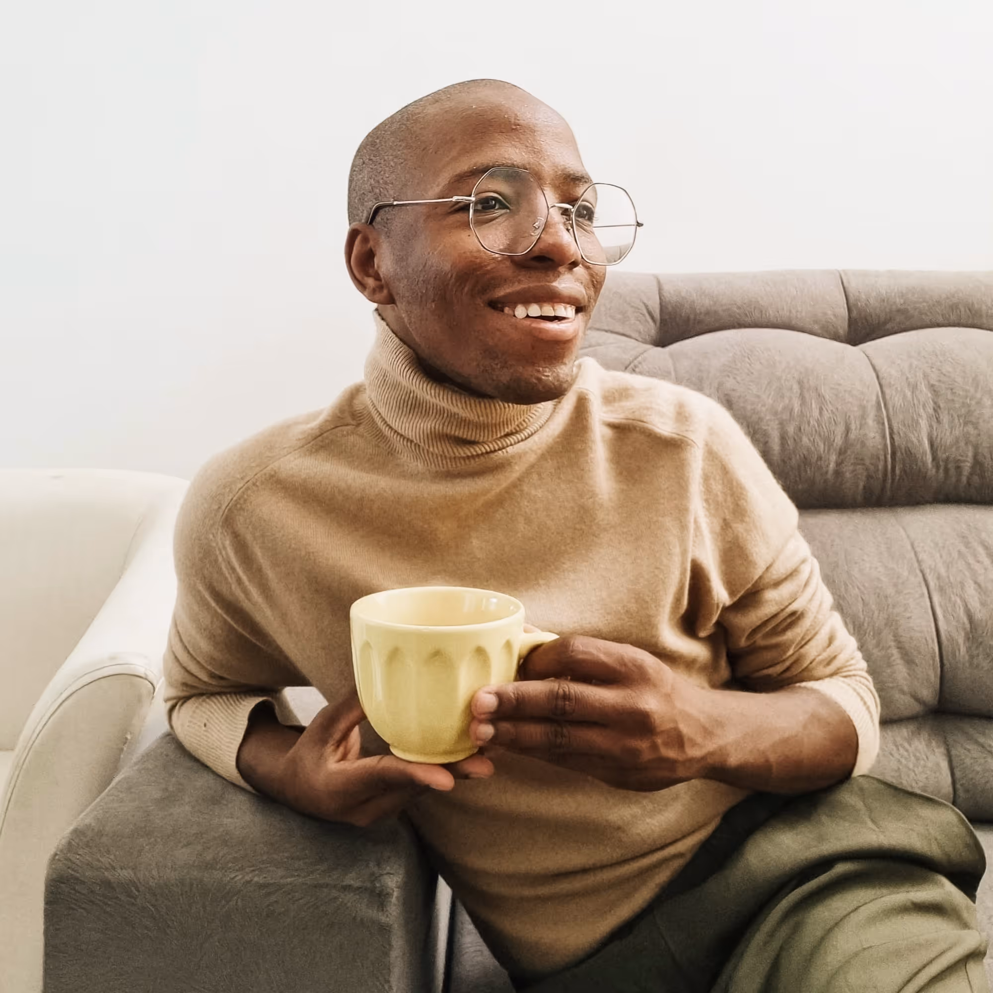 Smiling person in glasses and beige turtleneck sitting on a grey couch holding a yellow mug.