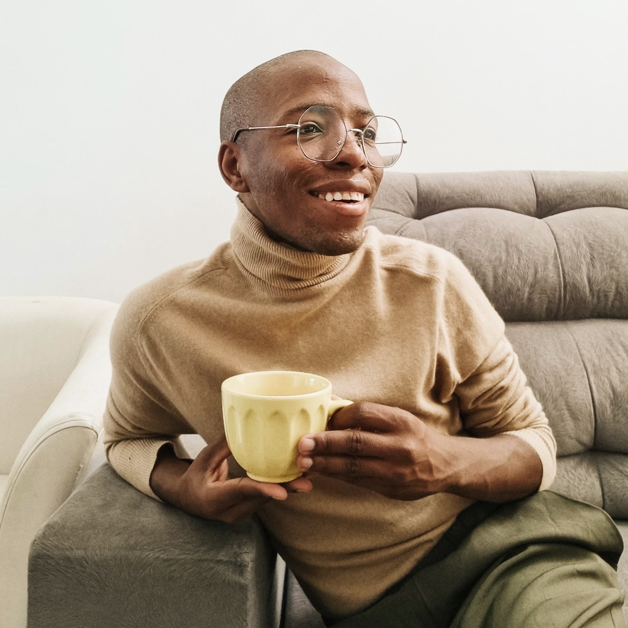 Smiling man with glasses wearing a beige turtleneck sitting on a gray couch holding a yellow mug.