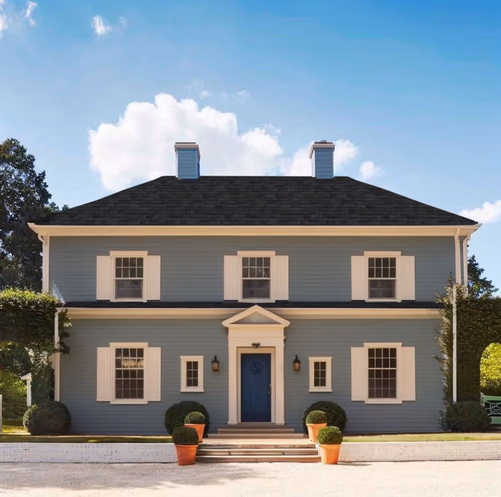 Two-story blue house with white shutters, black roof, a central blue door, and potted plants by the entrance.