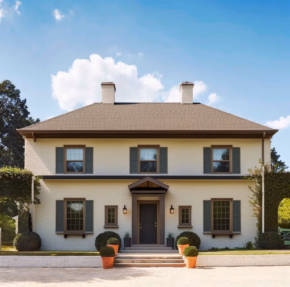 Two-story beige house with a brown door, six windows with green shutters, and four potted plants at the entrance.