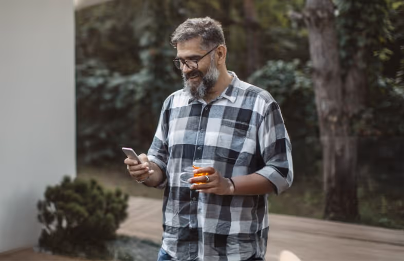 Man in a black and white checkered shirt holding a glass of orange drink in one hand and looking at his smartphone outdoors.