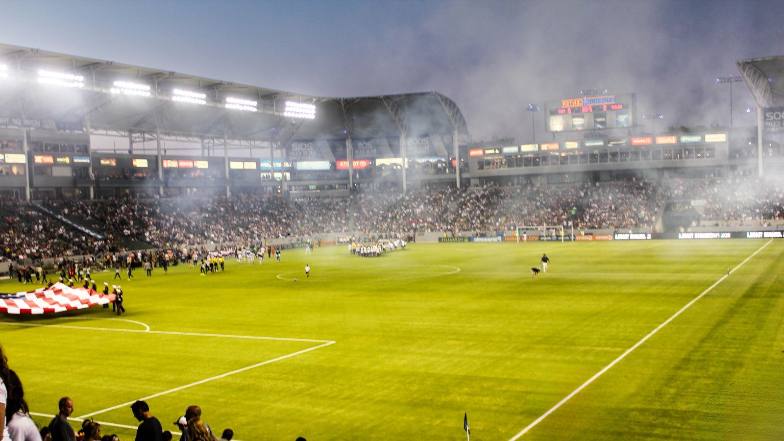 Football stadium packed with fans before kickoff, teams lined up on the pitch under floodlights, creating an intense matchday atmosphere during a competitive evening game.