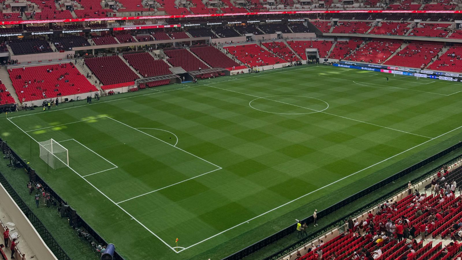 Wide-angle view of a professional football stadium before kickoff, showing a pristine green pitch, empty goalposts, red seating sections and spectators gradually taking their seats.