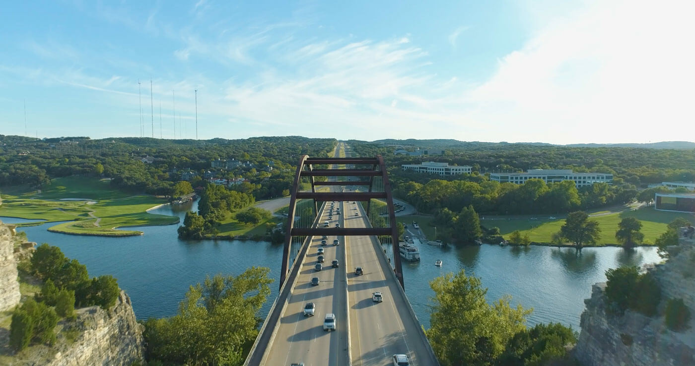 Aerial view of a busy Pennybacker bridge over a river, surrounded by lush greenery and a golf course, with a blue sky and wispy clouds.