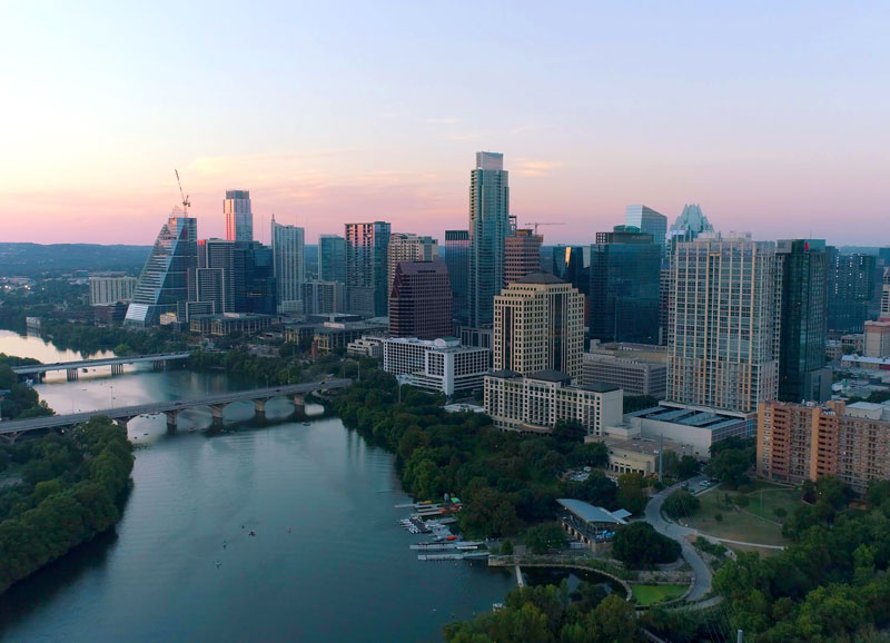 Aerial view of Austin cityscape with skyscrapers, bridge, and river, showcasing Texas capital's urban landscape at sunset with lush greenery and modern architecture.