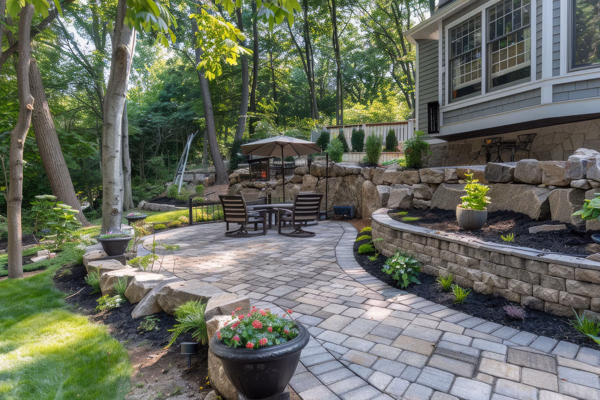 Backyard patio with stone pavers, curved retaining walls, outdoor seating under an umbrella, and surrounding greenery.