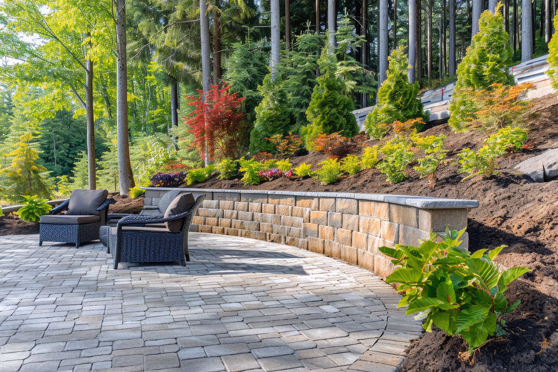 Outdoor patio with wicker chairs and ottoman on grey stone pavers next to curved retaining wall and landscaped plants.