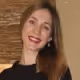 Smiling woman with straight brown hair wearing a black top in front of a wooden background.