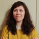 Woman with long brown hair wearing a yellow sweater against a plain background.