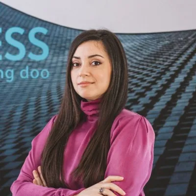 Woman with long dark hair wearing a purple top, standing with arms crossed in front of a blurred background with blue text.