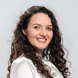 Smiling woman with long curly hair wearing a white top and pink blazer against a light gray background.