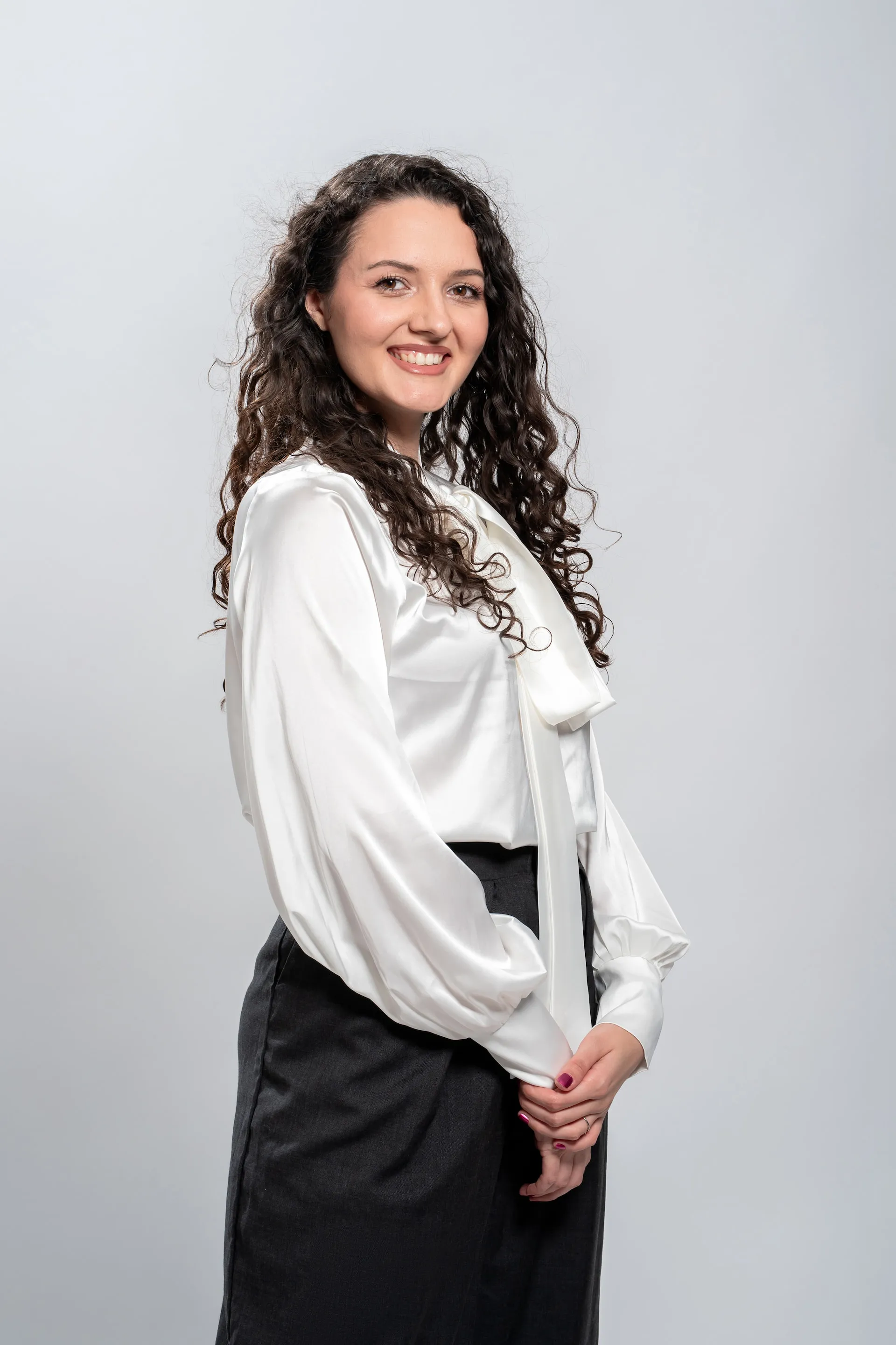 Smiling woman with curly hair wearing a pink blazer and white turtleneck standing in front of a whiteboard.