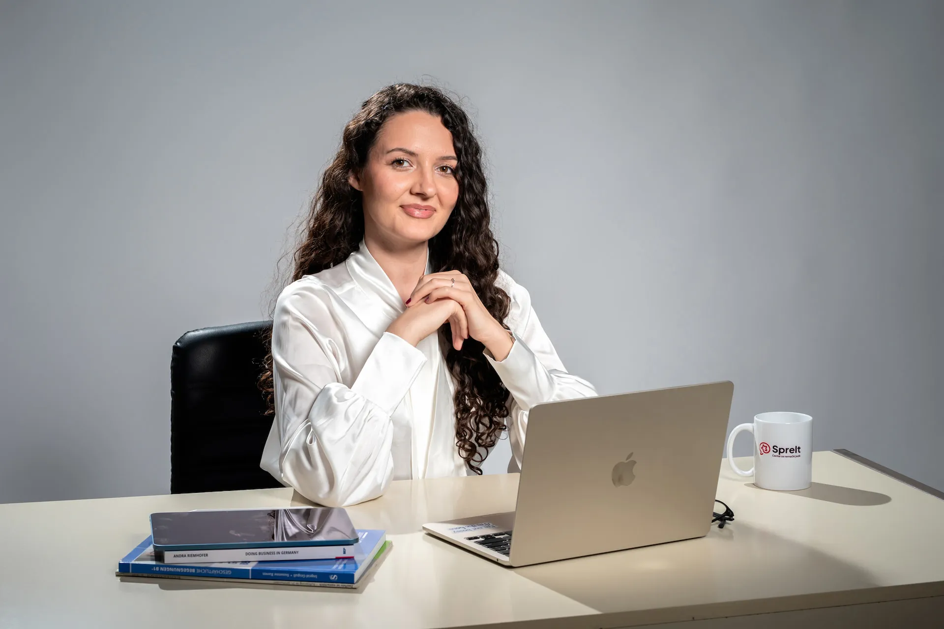 Smiling woman with curly hair wearing headphones and a navy blazer, looking at a laptop screen in a cozy indoor setting.