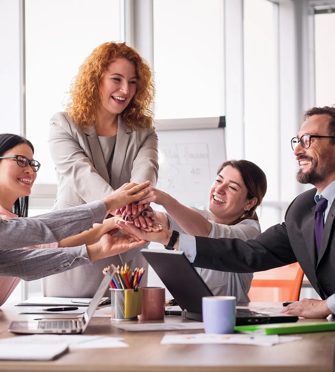Four diverse business colleagues putting hands together in a teamwork gesture around a desk with laptops and office supplies.