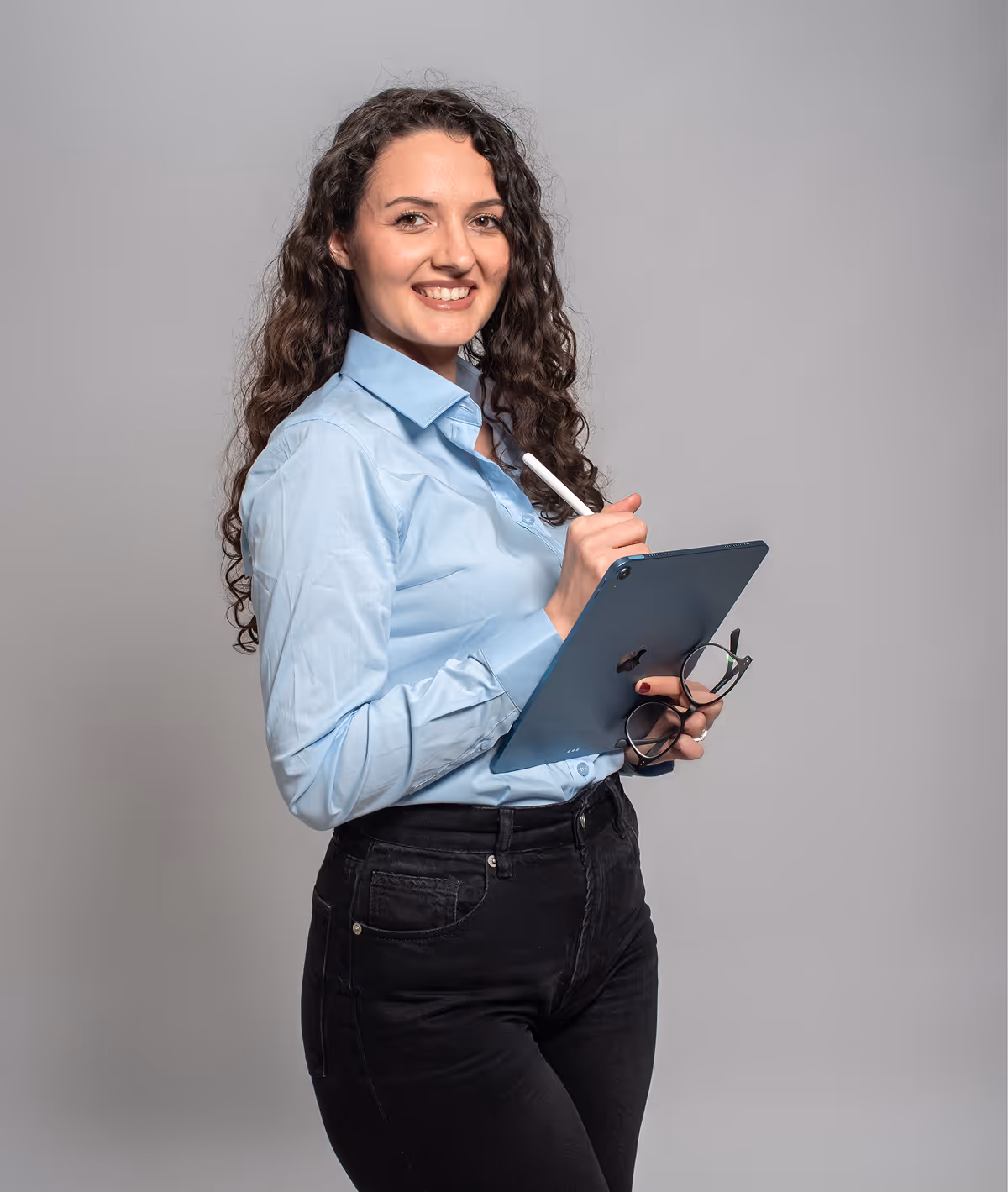 Smiling woman with curly hair wearing a pink blazer and white top standing in front of a whiteboard.