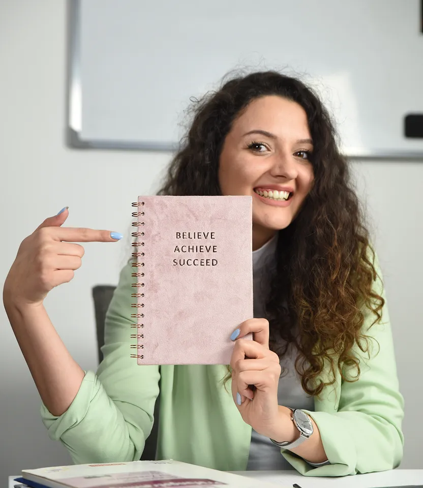 Overhead view of a person with curly hair sitting at a desk using a laptop and pointing at a planner with colored markings, with a tablet and headphones nearby.