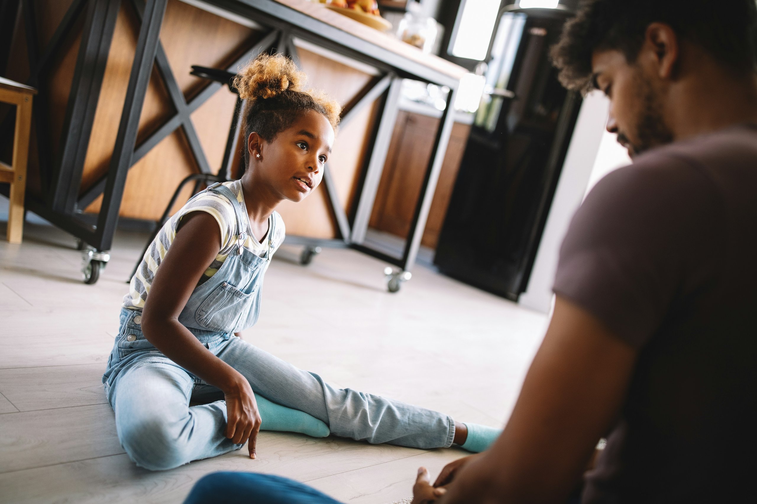 Young girl in denim overalls sitting on floor talking with adult