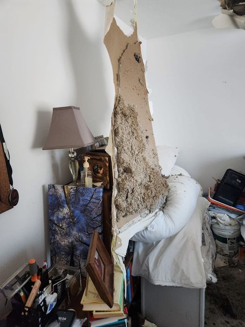 Severely damaged bedroom wall with large plaster and drywall debris falling onto a bed and cluttered nightstand.